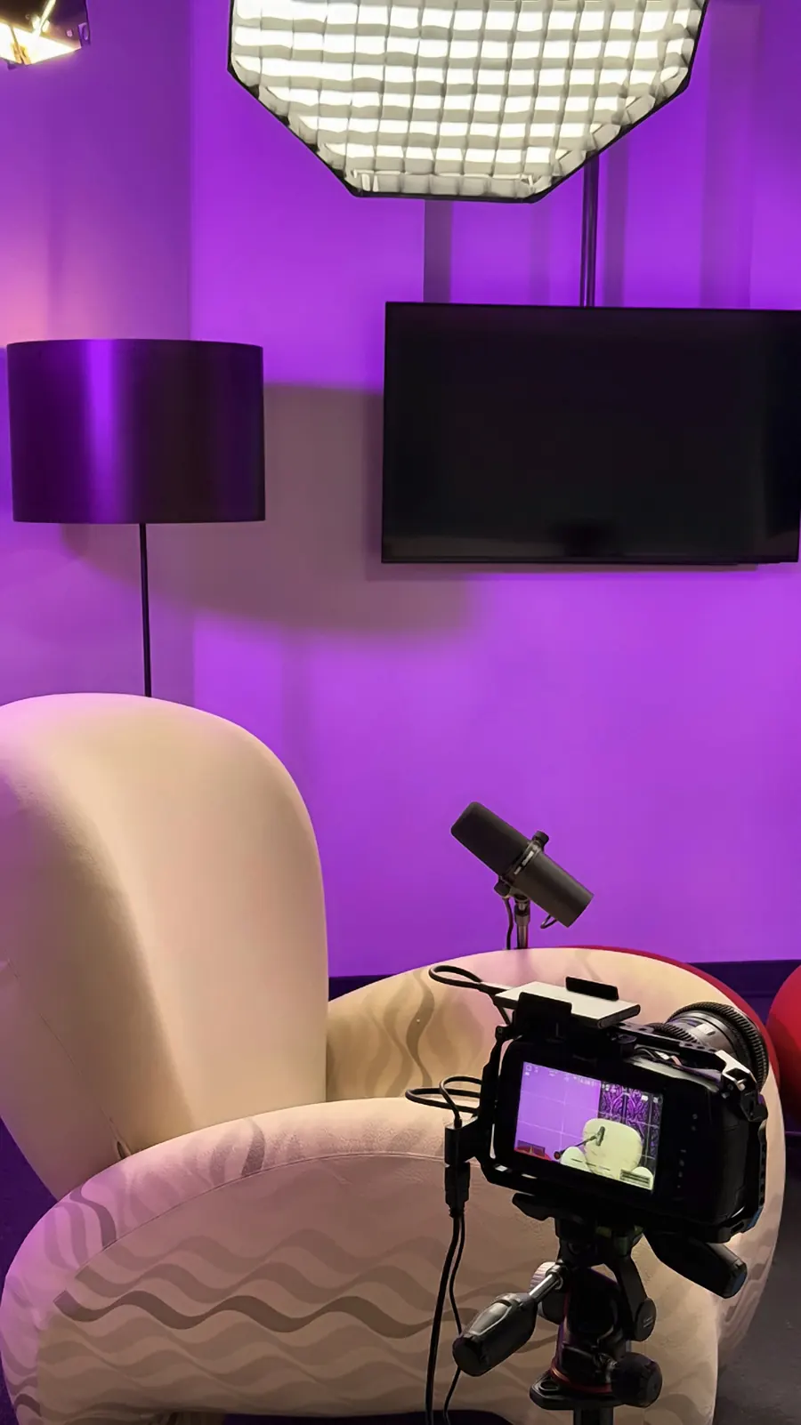Το Video Podcast Studio με μωβ φωτισμό: πολυθρόνα, μικρόφωνο, τηλεόραση στον τοίχο και κάμερα σε τρίποδο